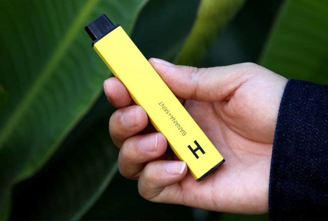 Person Holding Yellow Vape with Leafy Background 1
